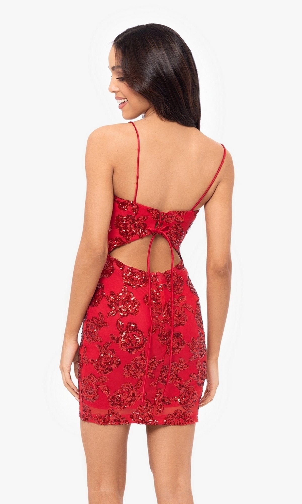 Woman wearing the Cut-Out Short Red Glitter Hoco Dress 4971BN, featuring sparkling details, thin straps, and a lace-up back, standing and looking to the side.